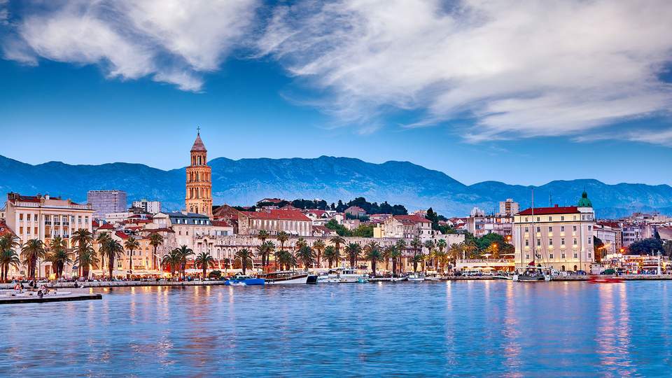 Man sieht vom Meer aus schauend einen Ausschnitt einer Stadt mit vor den Bergen und mit Palmen und alten Häusern