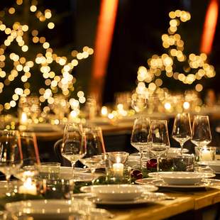 Festlich gedeckte Tafel für eine Weihnachtsfeier mit elegant arrangierten Gläsern und Tellern, umgeben von warmem Kerzenschein und verschwommenen, leuchtenden Weihnachtslichtern im Hintergrund in Form von Sternen und Bäumen.