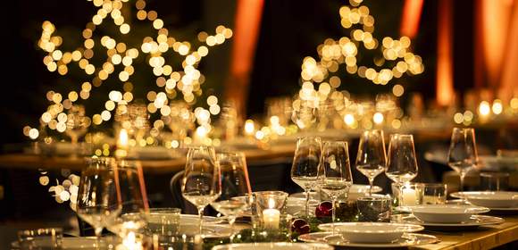 Festlich gedeckte Tafel für eine Weihnachtsfeier mit elegant arrangierten Gläsern und Tellern, umgeben von warmem Kerzenschein und verschwommenen, leuchtenden Weihnachtslichtern im Hintergrund in Form von Sternen und Bäumen.