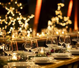 Festlich gedeckte Tafel für eine Weihnachtsfeier mit elegant arrangierten Gläsern und Tellern, umgeben von warmem Kerzenschein und verschwommenen, leuchtenden Weihnachtslichtern im Hintergrund in Form von Sternen und Bäumen.
