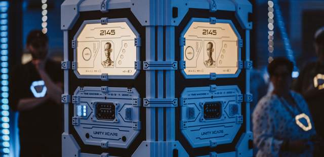 Eine futuristische blaue Zeitmaschine mit der Aufschrift 2145 und Unity Xcape, beleuchtet von blauem Ambientelicht, zeigen beleuchtete Panels mit KI-Gesicht und Time Machine Beschriftung im mobilen Escape Room