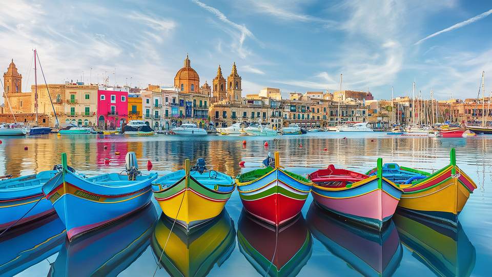 Man sieht eine Reihe bunter Fischerboote auf dem Wasser schwimmen und im Hintergrund eine Häuserreihe und einige Kirchtürme