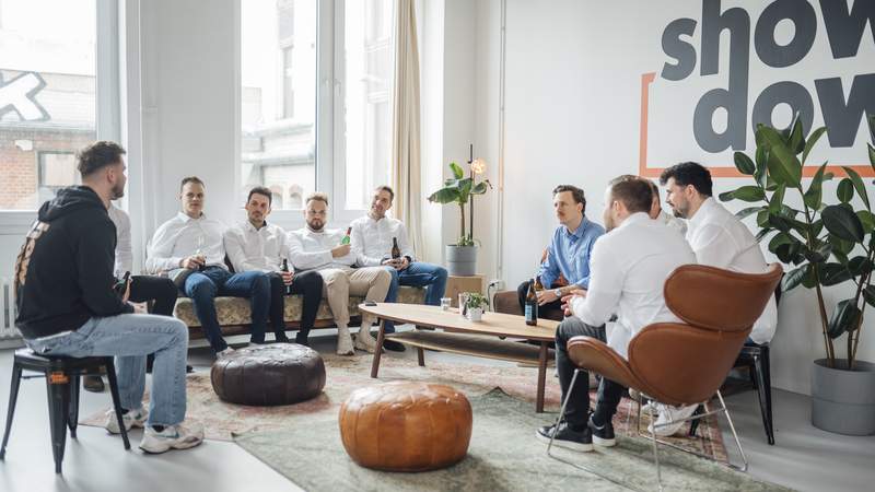 Auf dem Bild sieht man eine Gruppe von Männern in Hemden, die sich unseren Showdown von unserem Game Master erklären lassen. Sie sitzen alle auf Couches und Stühlen.