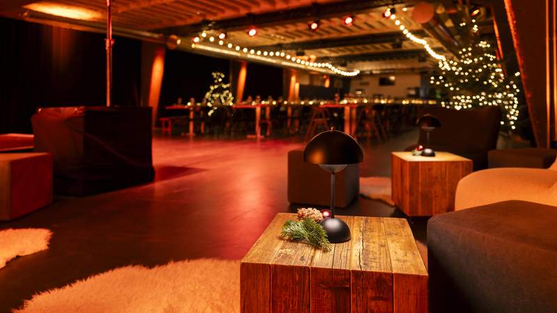 Weihnachtsfeier "Weihnachtlicher Orient" Gemütlich beleuchteter Loungebereich mit modernen Sesseln und rustikalen Holztischen, dekoriert mit Tannenzweigen und kleinen Lampen. Im Hintergrund sind festlich gedeckte Tafeln und Weihnachtsbäume mit Lichterketten zu sehen, die dem Raum eine warme