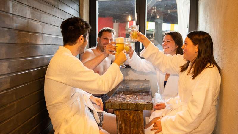 Vier Menschen in Bademänteln bekleidet sitzen beisammen und stoßen mit gefüllten Gläsern an