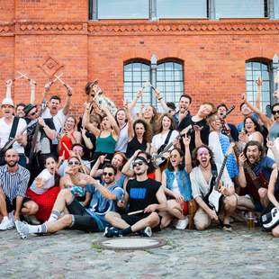 Große Gruppe spielt bei einem Teamevent als Rockband gemeinsam Instrumente, jubelt vor Backsteinfassade und erlebt intensives Teambuilding ohne Vorkenntnisse.