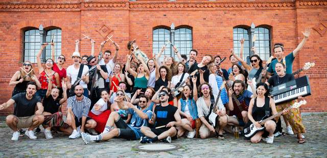 Große Gruppe spielt bei einem Teamevent als Rockband gemeinsam Instrumente, jubelt vor Backsteinfassade und erlebt intensives Teambuilding ohne Vorkenntnisse.
