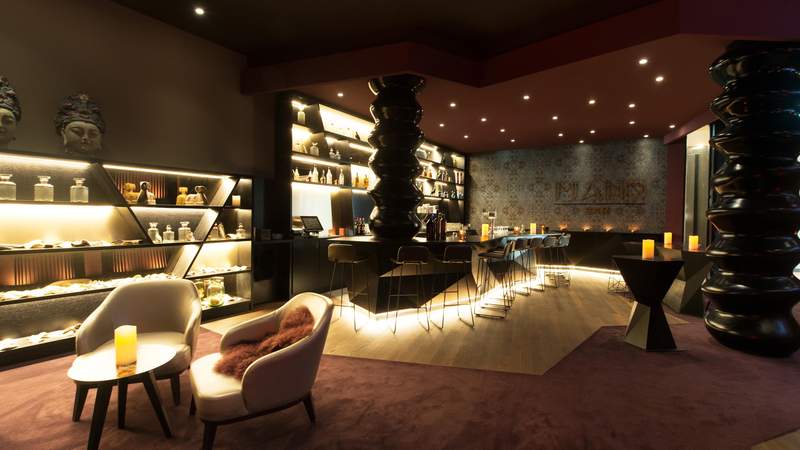 Gemütlich beleuchtete Piano Bar mit Sesseln im Vordergrund und der Bar mit Barhockern und schwarzen Säulen im Hintergrund.