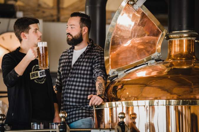 Zwei Herren vor einem Bierkessel, einer hält ein Glas Bier in der Hand.