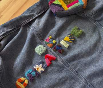 Jeansjacke auf Holzhintergrund mit bunten Punch-Needle-Buchstaben und geometrischen Patches